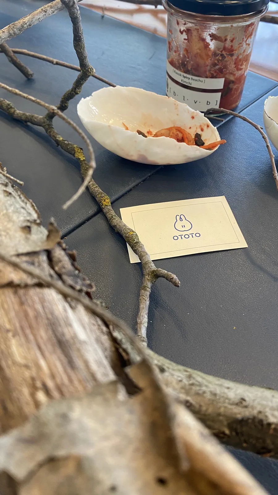 A weathered wooden log and bare twigs on the grey table next to a shell-shaped dish of kimchi, the jar of BLVB kimchi behind and the OTOTO card