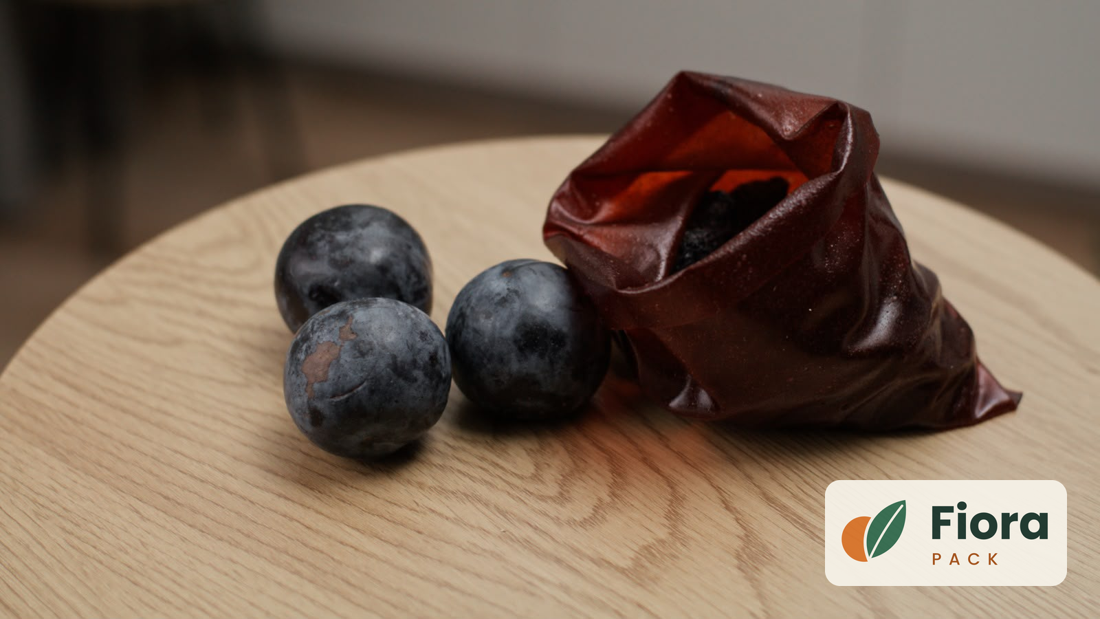 Three dark plums beside an open, translucent amber pouch of biodegradable food film on a light wooden tabletop — packaging by Fiora Pack.