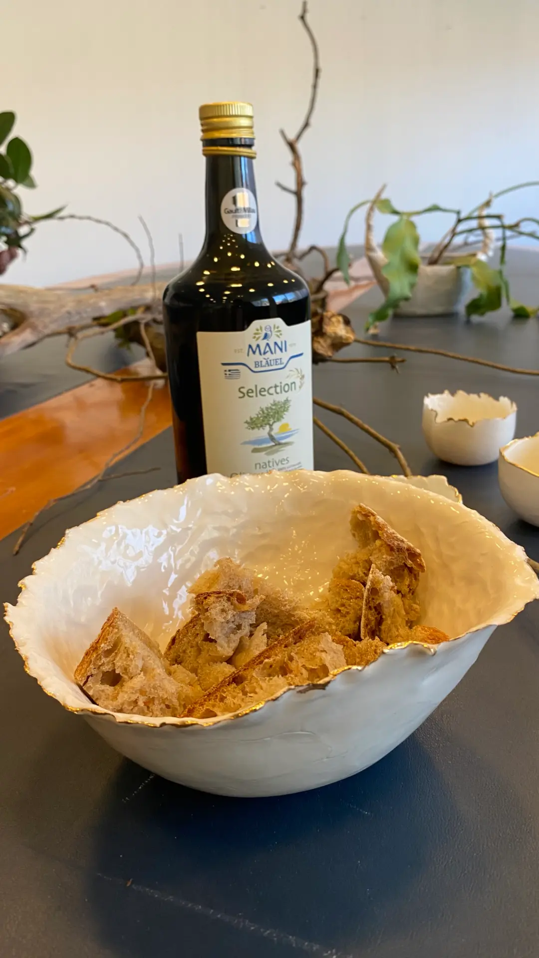 MANI Bläuel olive oil standing beside a torn porcelain bowl with gilded rim, filled with broken sourdough, branches and small vessels behind