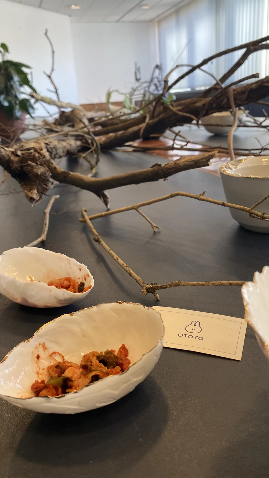 Two gold-rimmed shell-shaped porcelain bowls of kimchi on the table in soft daylight, dry branches crossing beside the OTOTO card