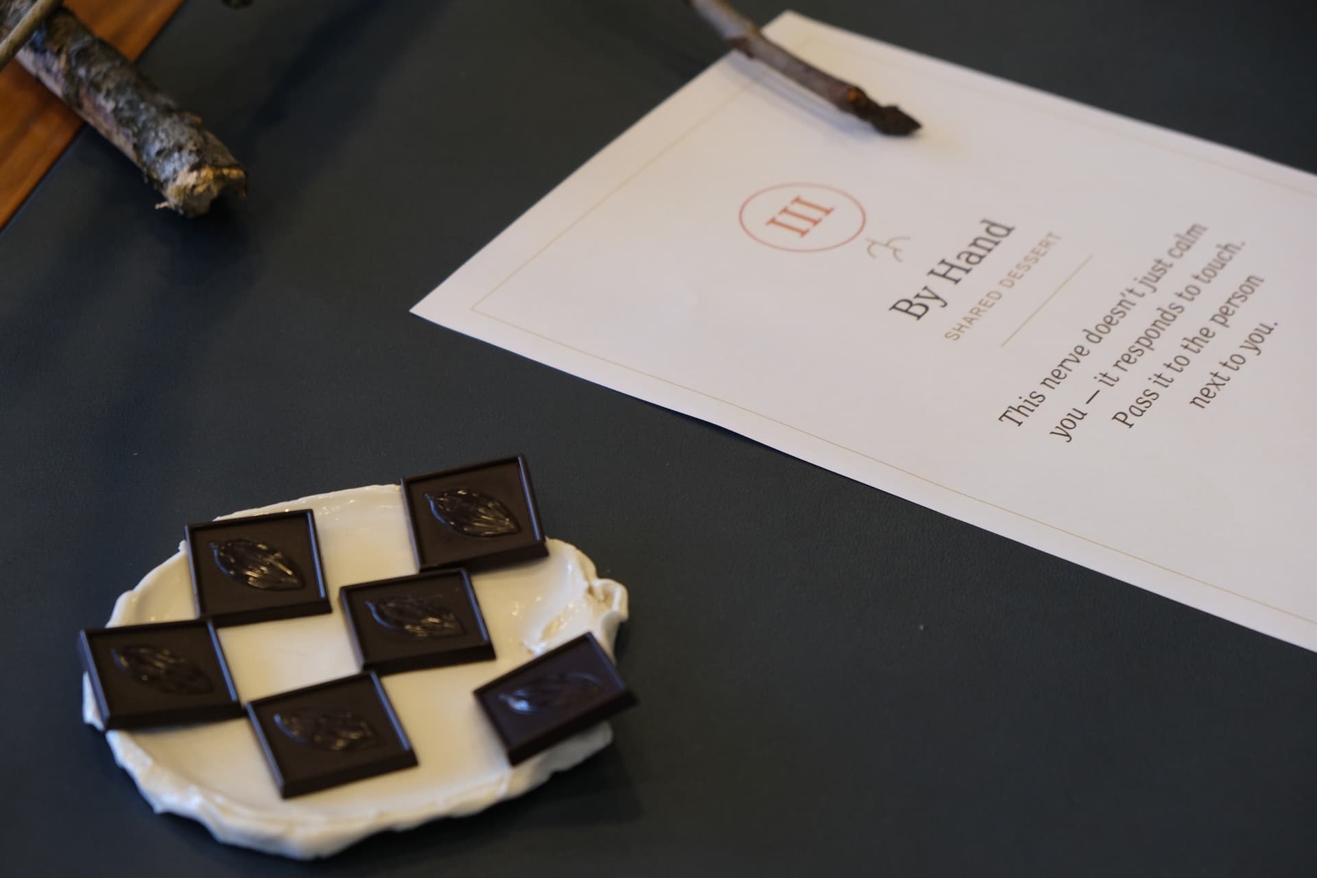 The printed card for station III, By Hand — Shared Dessert, resting on the table beside chocolate squares on porcelain