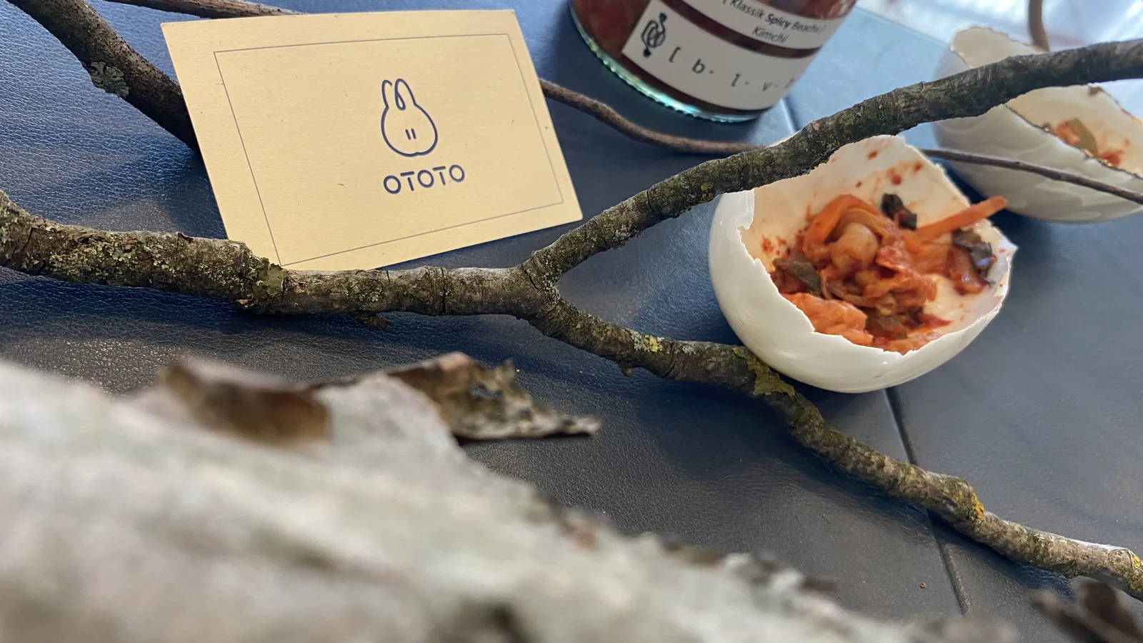 Wide view of the fermentation station: lichen-covered twigs, a porcelain bowl of kimchi and a jar of BLVB Klassik Spicy Baechu resting on the grey table with the OTOTO card