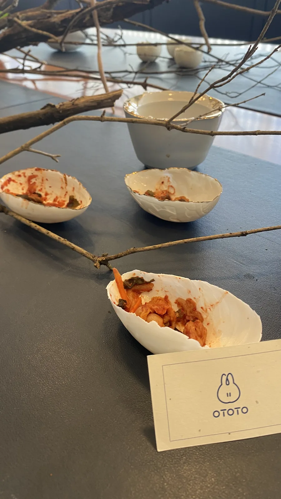 The fermentation station seen from above: kimchi in pale porcelain pinch bowls, a gold-rimmed cup, branches laid across the grey table and the OTOTO card