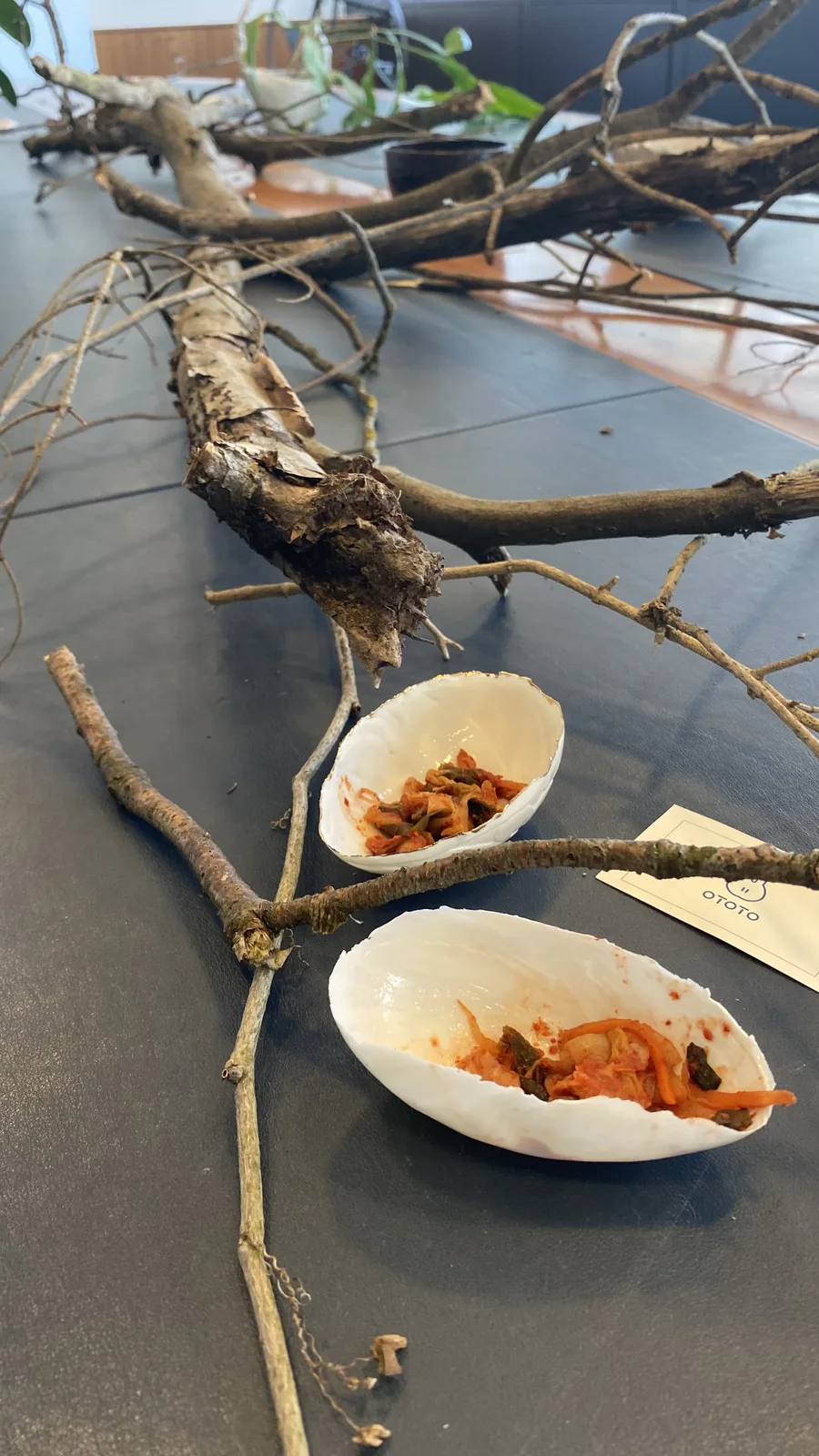 A long weathered branch laid the length of the dark table beside two shell-shaped bowls of kimchi and the OTOTO card