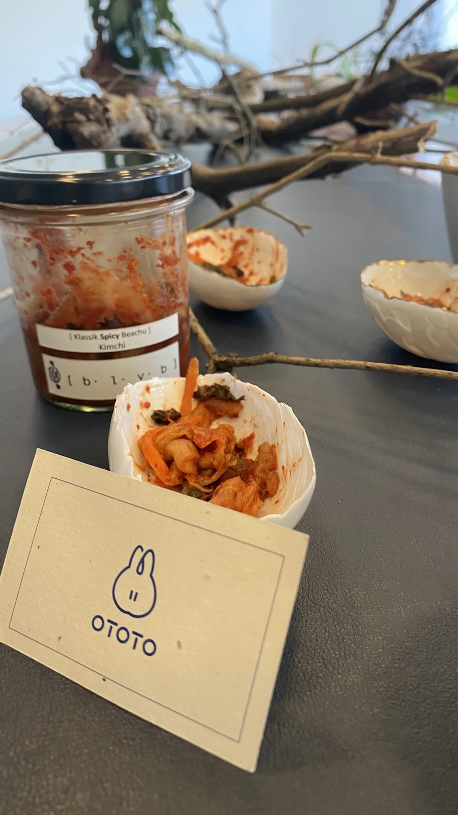 A glass jar of BLVB Klassik Spicy Baechu kimchi with a black lid standing behind a shell-shaped porcelain bowl of kimchi and the cream OTOTO card