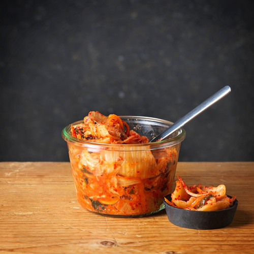 A jar of Blvb-blub KG kimchi with a serving spoon on a wooden table.