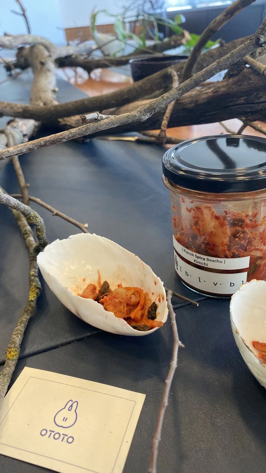 A shell-shaped porcelain bowl of kimchi among dry branches, with a jar of BLVB Klassik Spicy Baechu and the cream OTOTO card printed with a blue rabbit