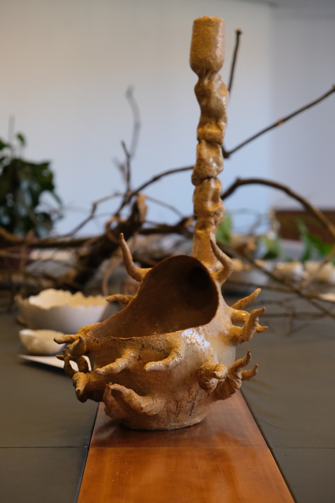 A sculptural ceramic vessel with root-like tendrils climbing into a tall stem, standing on the exhibition table among branches