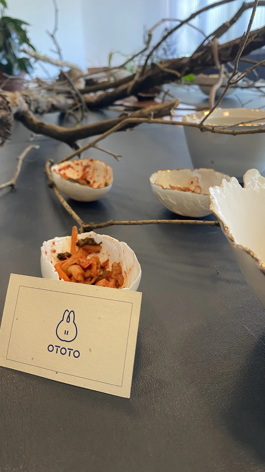 Jagged-edged white porcelain dishes holding kimchi, photographed at an angle across tangled bare branches on the dark table, the OTOTO card in the foreground