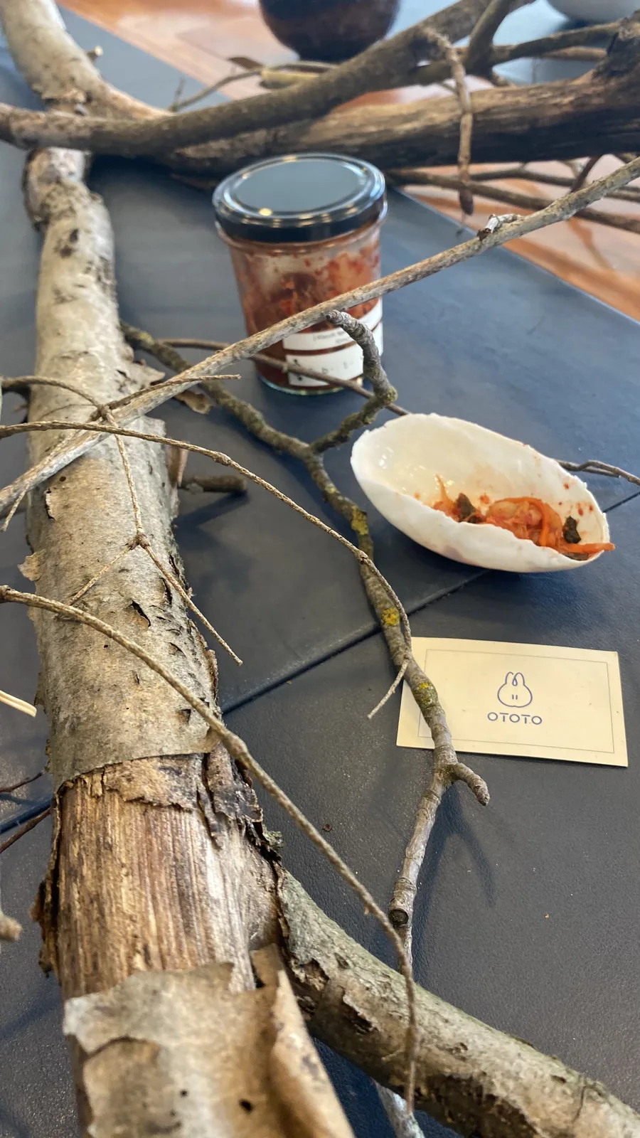 A thick branch with peeling bark stretching across the dark table beside a shell-shaped dish of kimchi, smaller twigs and the OTOTO card