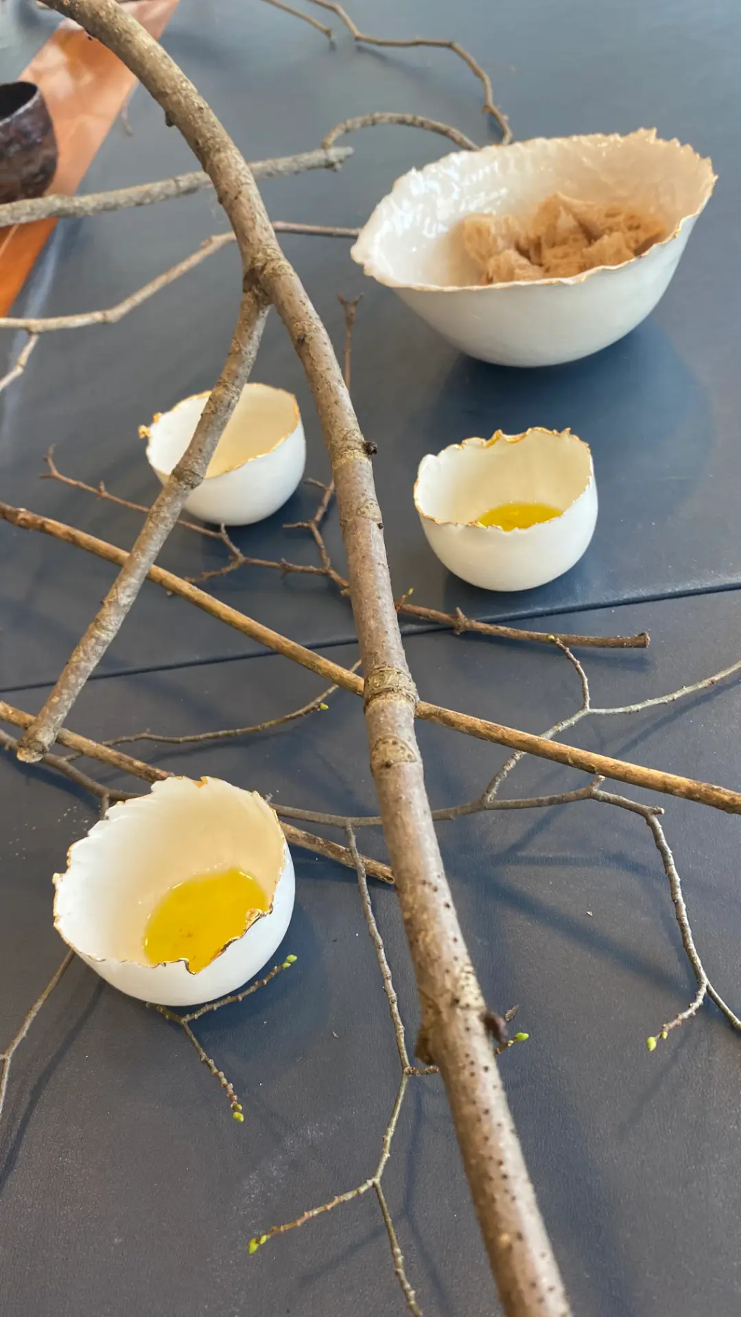 Small porcelain vessels with gilded rims holding pools of olive oil, set among bare branches crossing the dark table