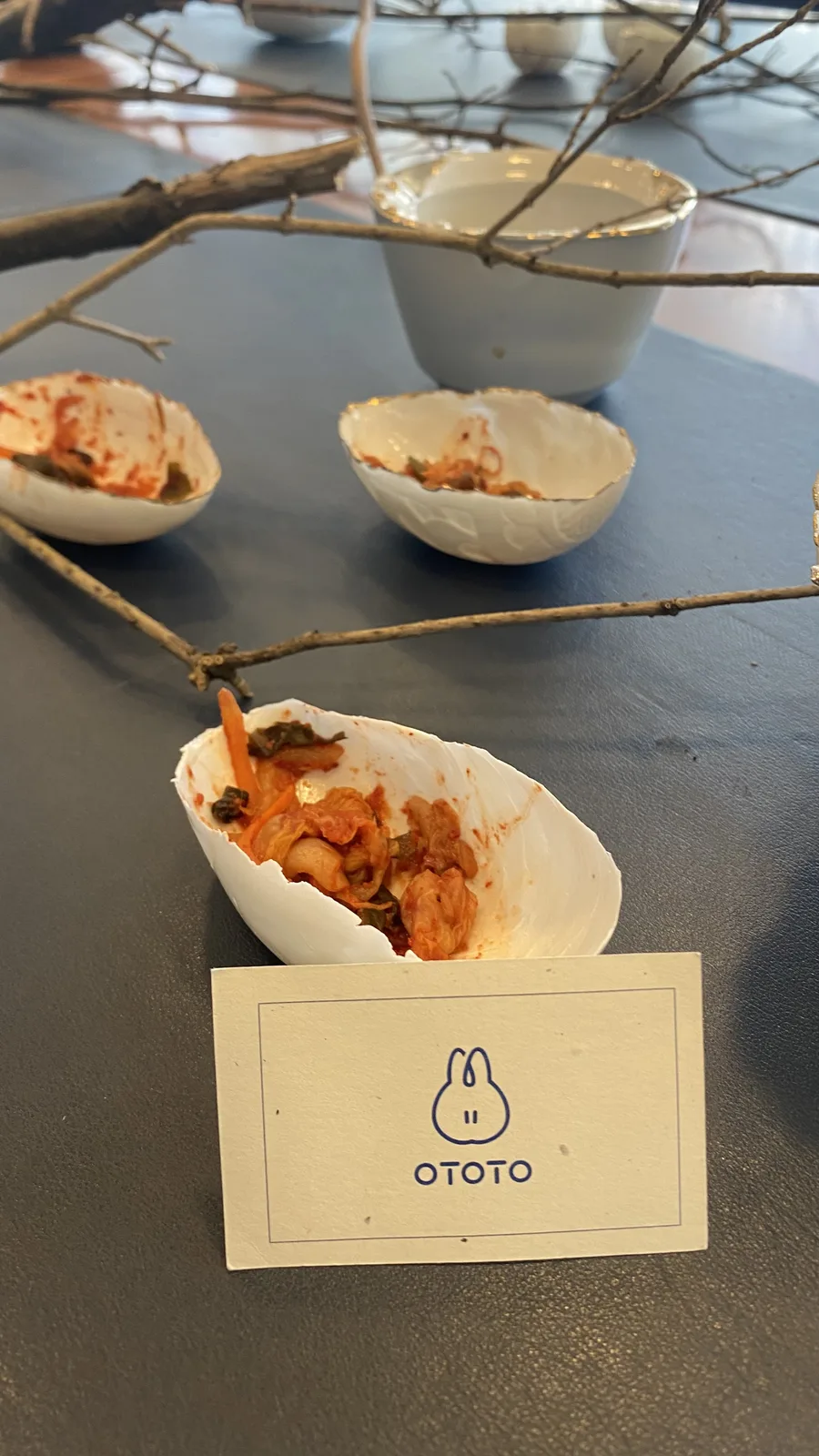 Small white eggshell-shaped porcelain bowls holding red kimchi on the dark exhibition table, a gold-rimmed cup and a cream card printed with a blue rabbit and the word OTOTO beside bare branches